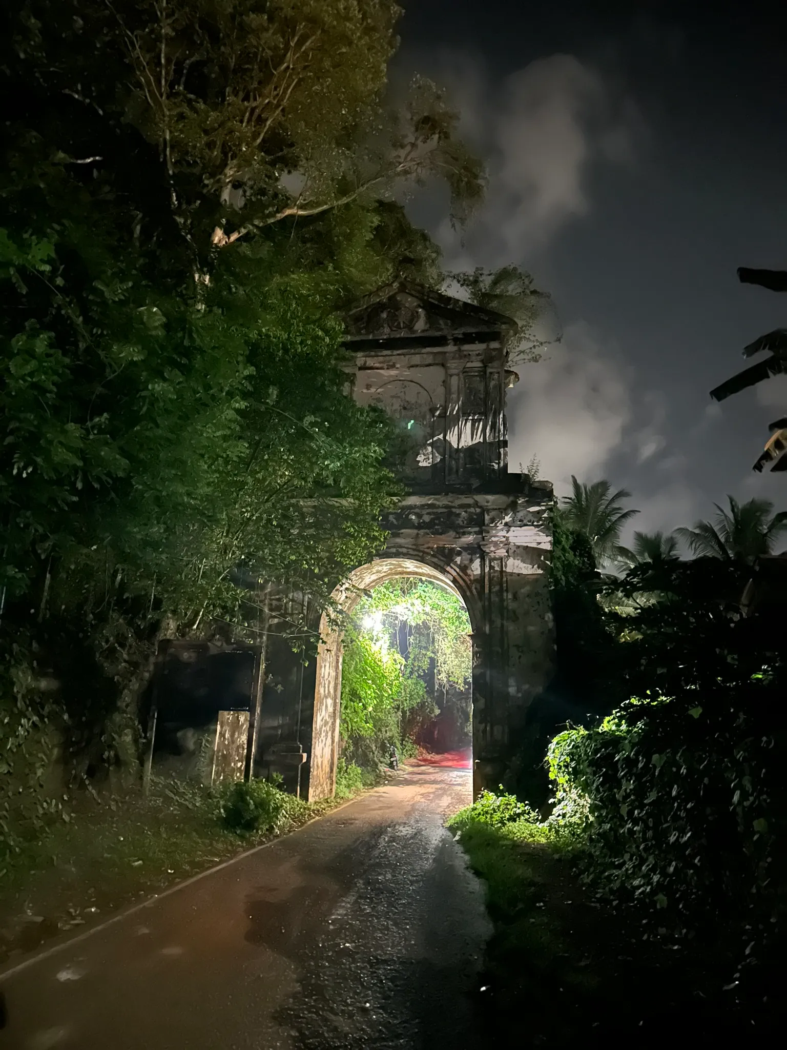 A ruined stone archway at night, overgrown with vines, a lit road passing through it.